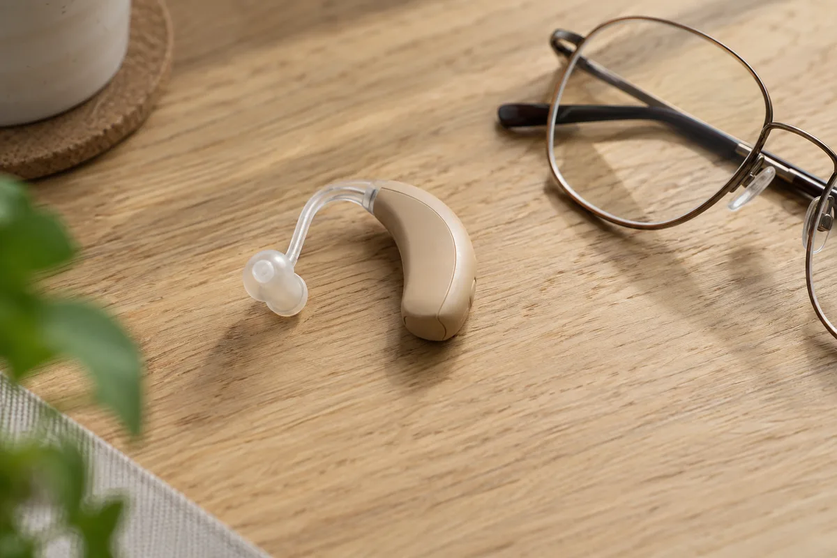 Petit appareil auditif beige uni posé à côté d&rsquo;une paire de lunettes sur une table en bois