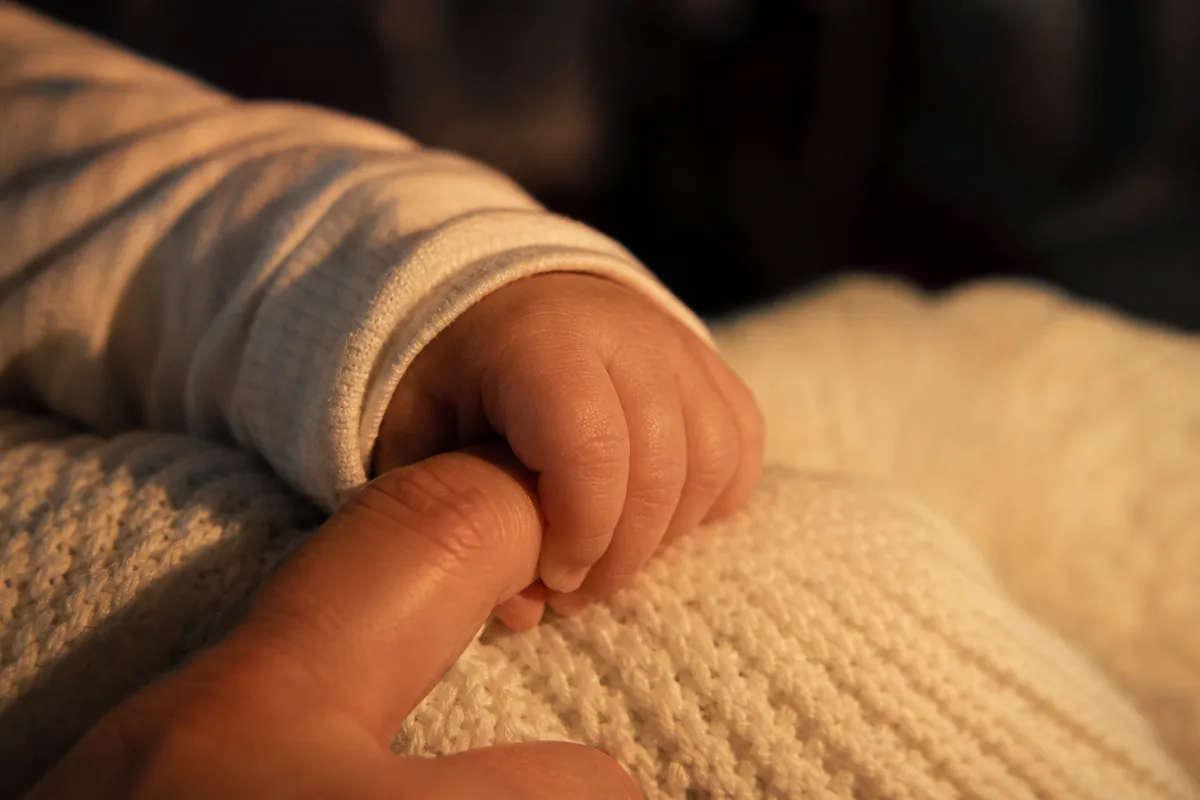 Petite main de bébé refermée sur le doigt d’un adulte, sur une couverture en maille claire, lumière tamisée de cabine
